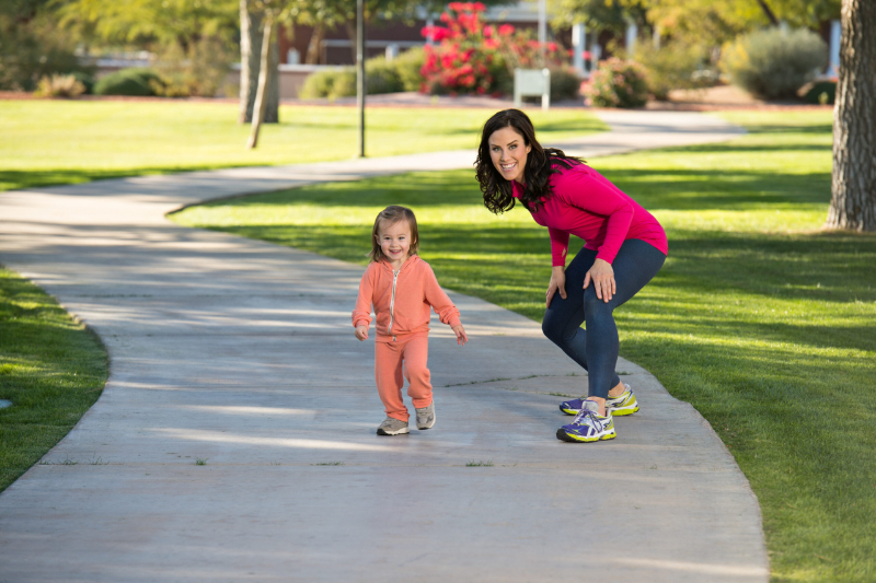 buy a home near a greenbelt, this is an image of Beautiful young mother and her daughter running in the neighborhood. They are on a sidewalk in a grassy greenbelt. The mother is playfully chasing her daughter.